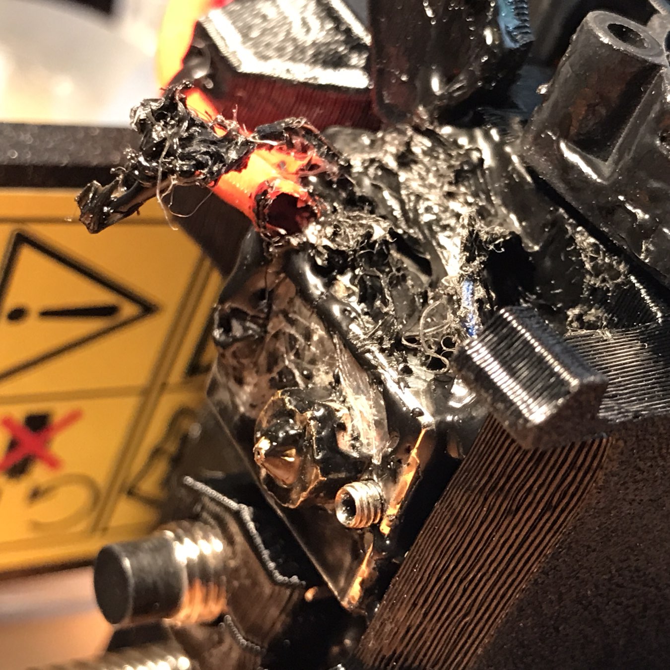 Auto-generated description: A 3D printer's hotend is shown with melted and burnt filament, alongside visible damage and a caution sign.